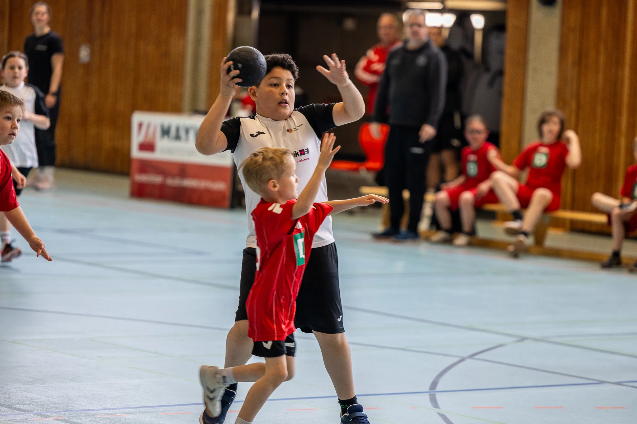 Ein Jugendlicher in schwarzem Trikot wirft einen Handball, während ein Kind in rotem Trikot ihm entgegenläuft. Im Hintergrund sitzen und stehen weitere Spieler und Zuschauer in einer Sporthalle.