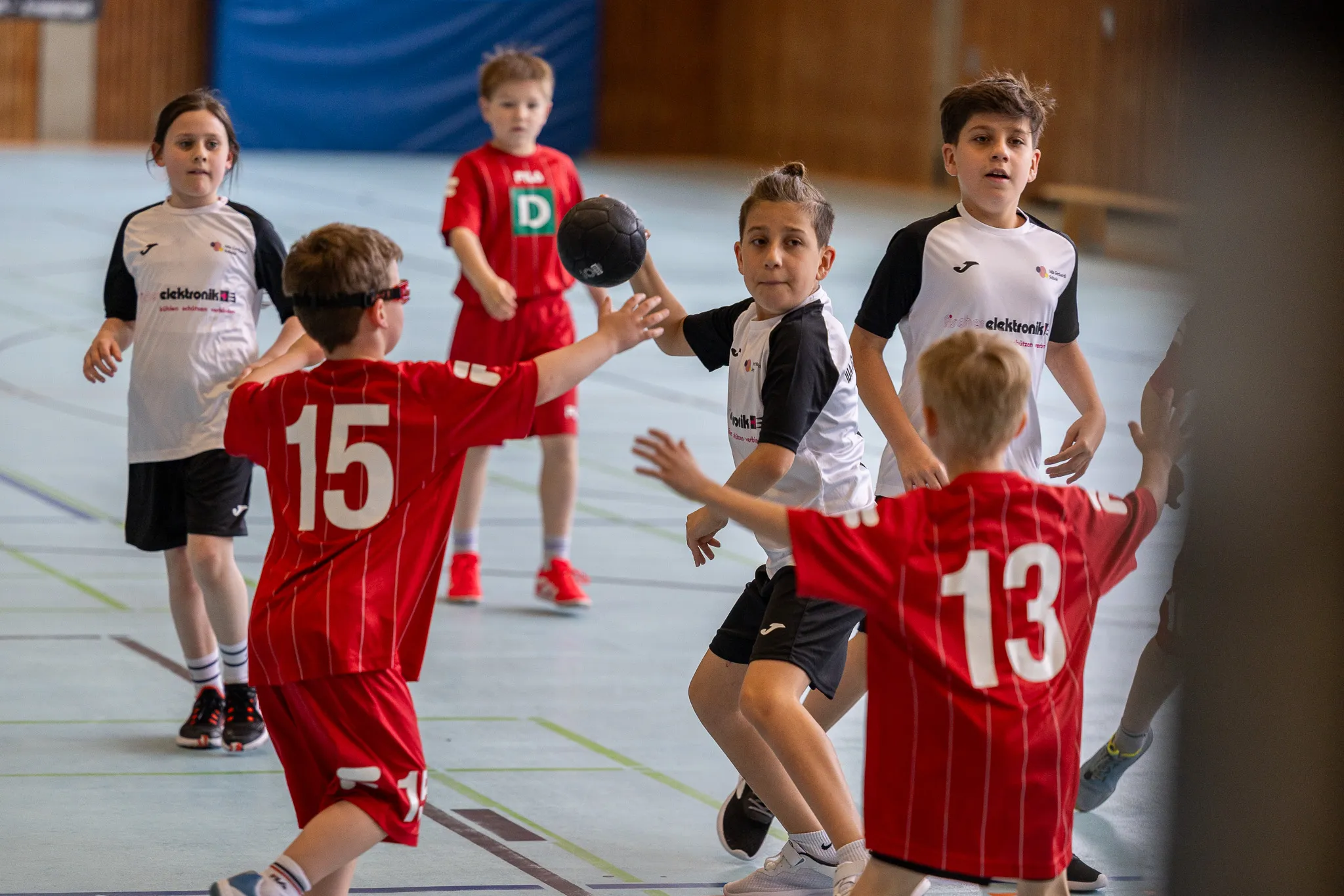 Zwei Jungen werfen sich in einer Sporthalle einen Ball zu, während andere Kinder im Hintergrund zusehen und mitspielen. Die Trikots tragen die Aufschrift eines Sponsors und Rückennummern.