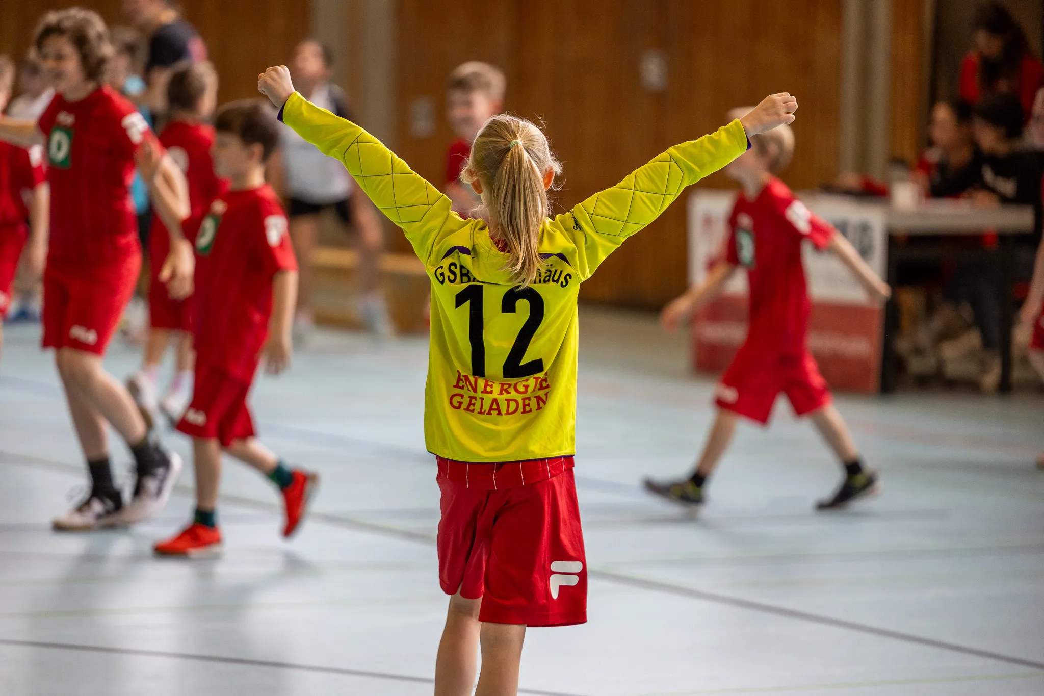 Junge im gelben Trikot mit der Nummer 12 und roten Shorts steht mit erhobenen Armen auf einem Hallenfußballfeld, umgeben von anderen Spielern in roten Trikots.