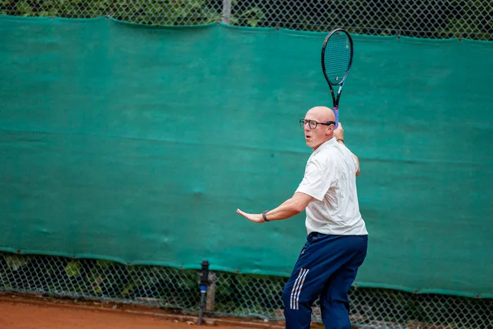 Ein Mann mit Brille und weißem Poloshirt schwingt einen Tennisschläger vor einem grünen Sichtschutz.