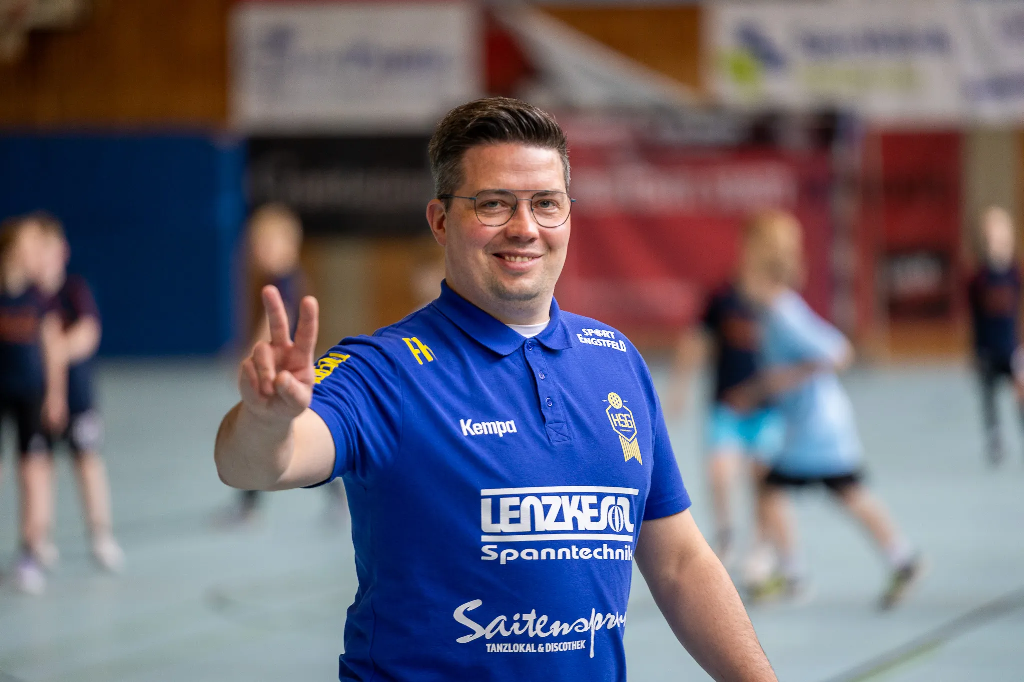 Ein Mann mit Brille und blauem Poloshirt zeigt beim Fotografieren die Victory-Geste. Im Hintergrund sind verschwommene Personen in einer Sporthalle zu sehen.