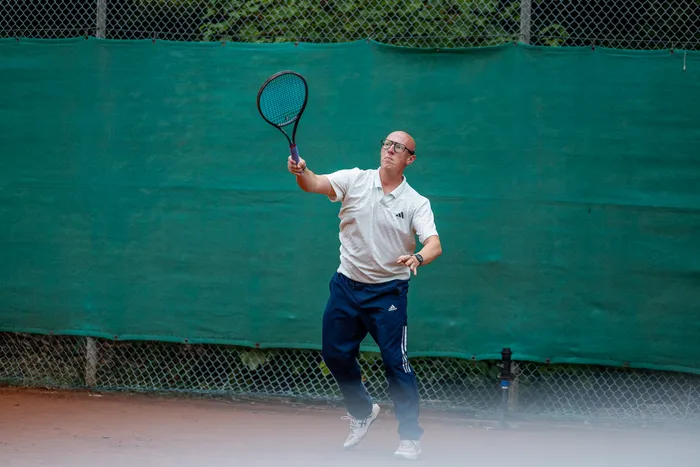Ein Mann in weißem Poloshirt und blauer Hose schwingt einen Tennisschläger auf einem Sandplatz vor einem grünen Zaun.
