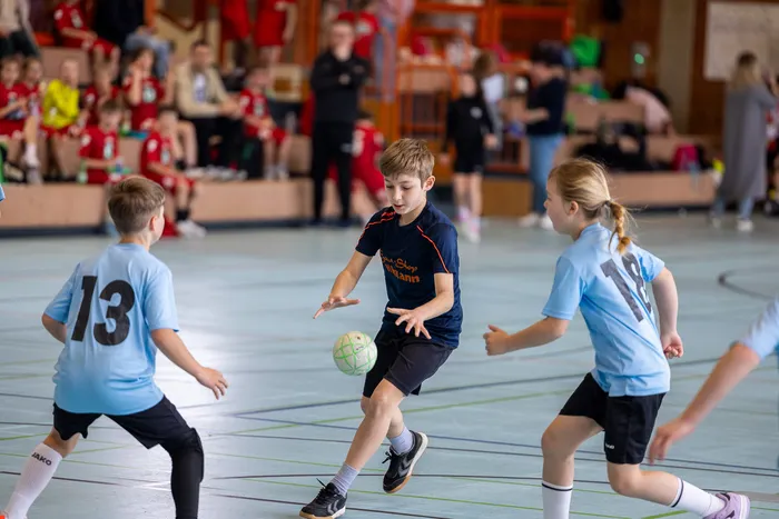 Drei Kinder spielen Hallenhandball. Ein Junge dribbelt den Ball, während zwei andere ihn verteidigen. Im Hintergrund sind Zuschauer auf einer Tribüne zu sehen.