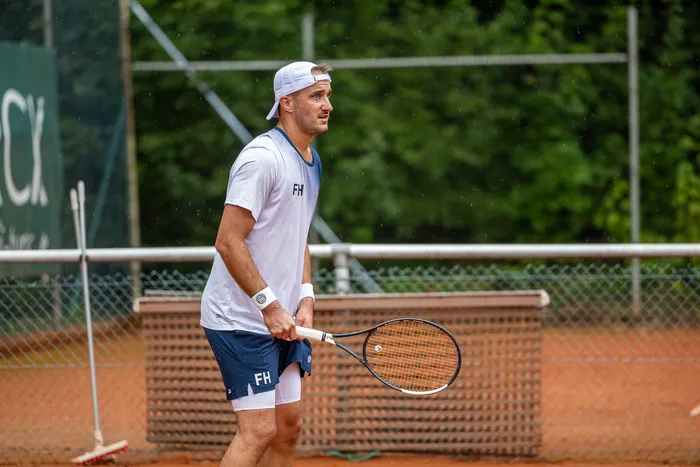 Ein Tennisspieler mit weißer Kappe und grauem T-Shirt mit der Aufschrift „FH“ steht mit einem Schläger auf einem Sandplatz.