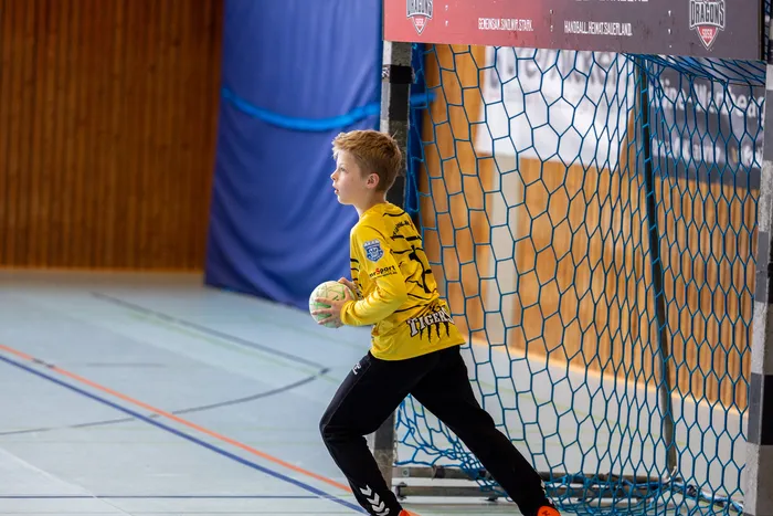 Ein Jugendlicher in Handballausrüstung steht vor einem Tor und hält einen Ball bereit. Im Hintergrund ist ein Handballfeld und Werbebanner zu sehen.