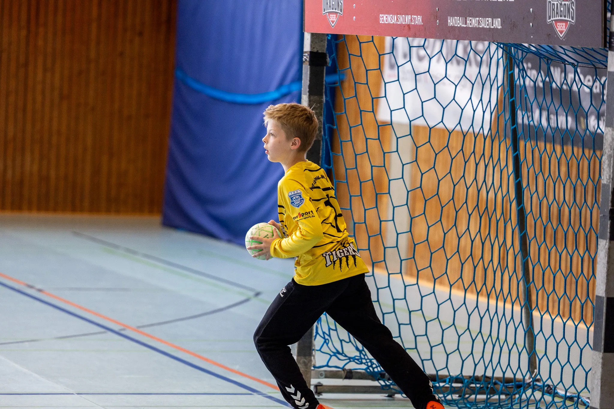Ein Jugendlicher in Handballausrüstung steht vor einem Tor und hält einen Ball bereit. Im Hintergrund ist ein Handballfeld und Werbebanner zu sehen.