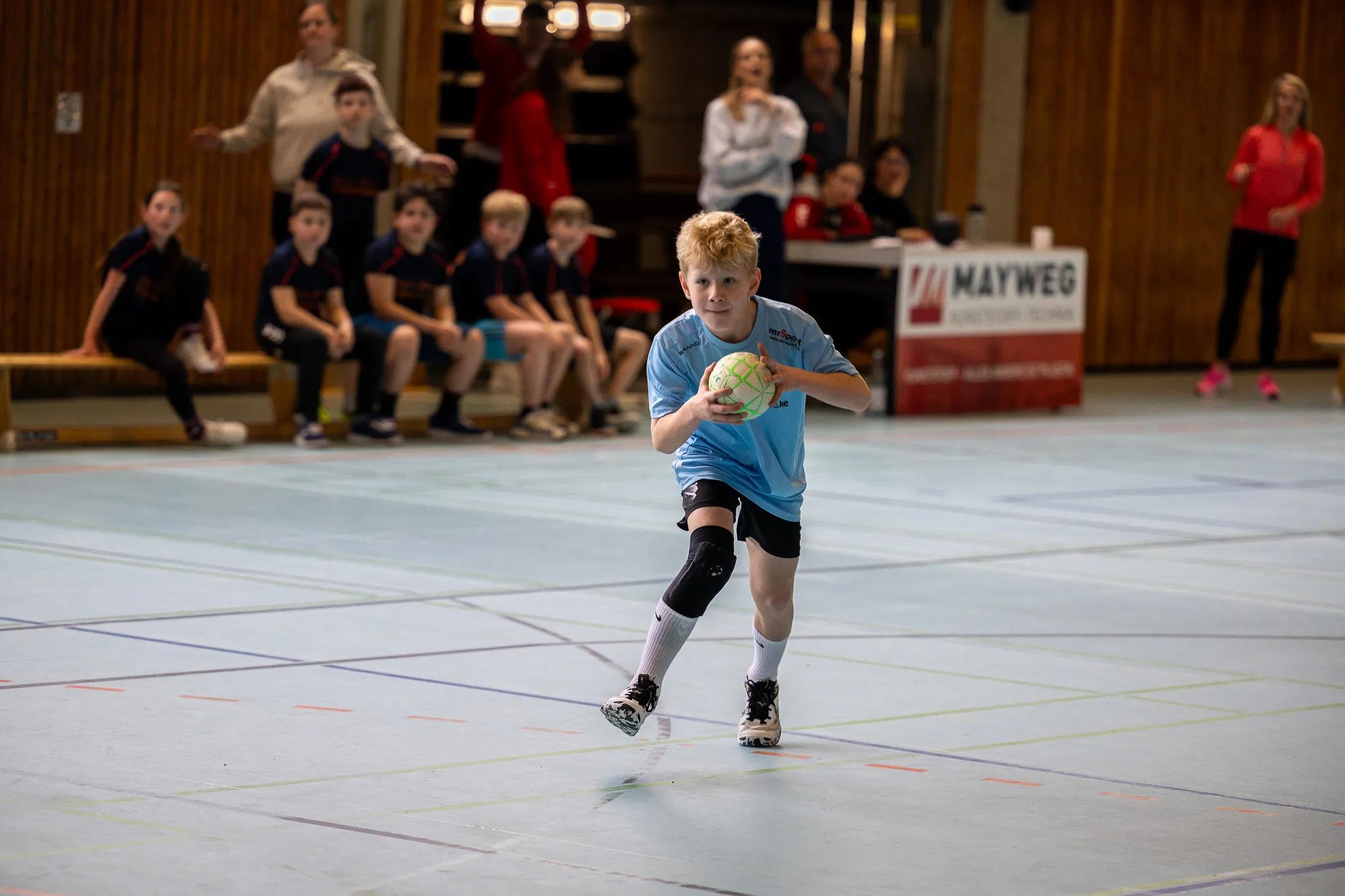 Ein Junge mit blonden Haaren dribbelt einen Handball in einer Sporthalle, während im Hintergrund Zuschauer sitzen und stehen.