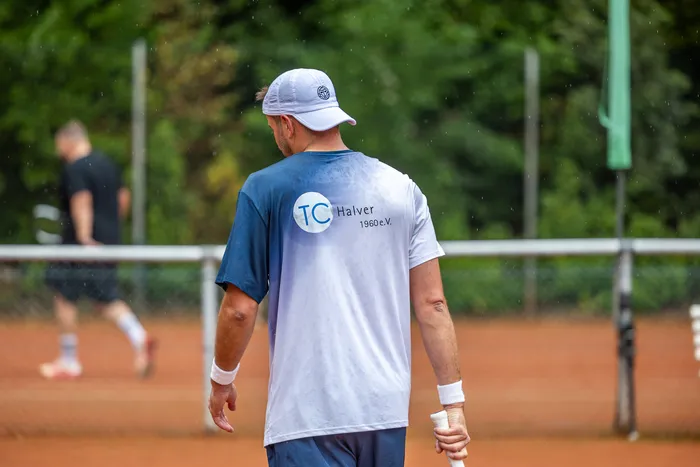 Ein Mann mit weißer Kappe steht mit dem Rücken zur Kamera auf einem Tennis-Sandplatz und trägt ein T-Shirt mit dem Aufdruck „TC Halver 1960 e.V.“.