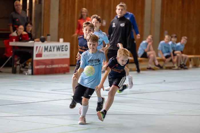Zwei Jungen in Handballtrikots sprinten während eines Spiels in einer Sporthalle. Im Hintergrund stehen weitere Kinder und ein Trainer.