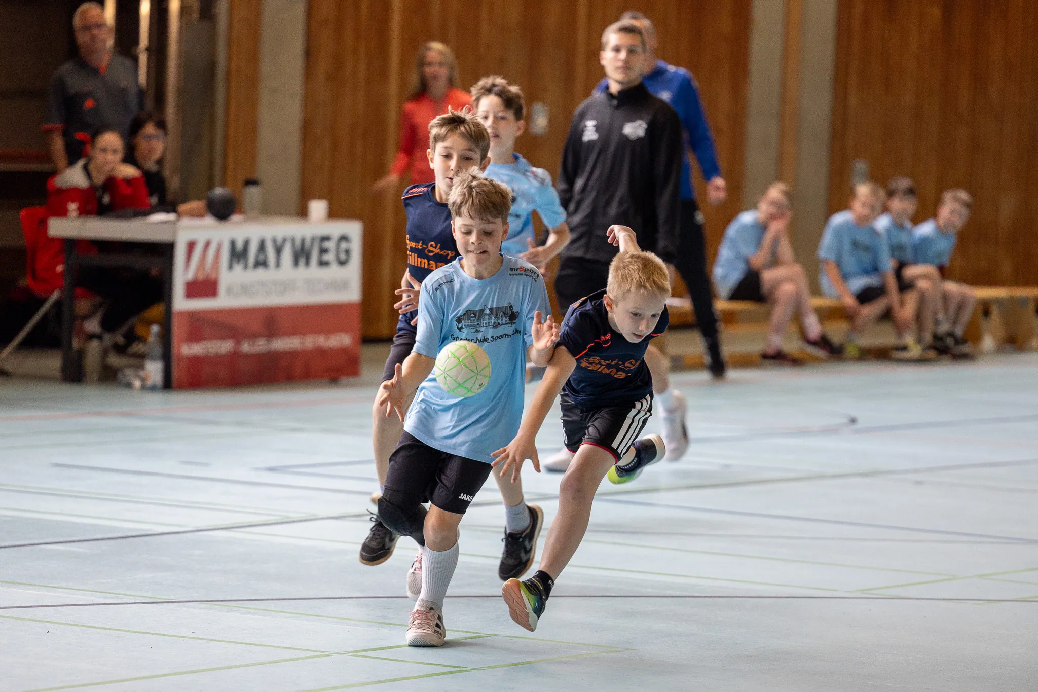Zwei Jungen in Handballtrikots sprinten während eines Spiels in einer Sporthalle. Im Hintergrund stehen weitere Kinder und ein Trainer.