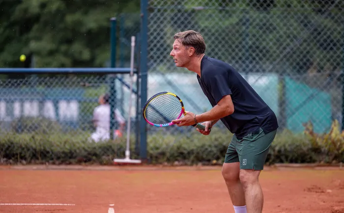 Ein Tennisspieler in dunklem T-Shirt und grünen Shorts steht in einer bereitstehenden Haltung mit dem Schläger auf einem Sandplatz.