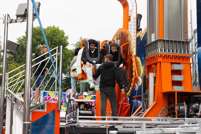Personen steigen in eine orangefarbene Fahrgeschäftskonstruktion, während andere zusehen. Im Hintergrund sind weitere Fahrgeschäfte und Besucher der Kirmes zu erkennen.