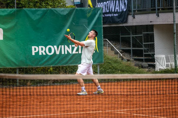 Ein Tennisspieler in weißer Kleidung schlägt einen gelben Ball auf einem Sandplatz vor einem grünen Banner.
