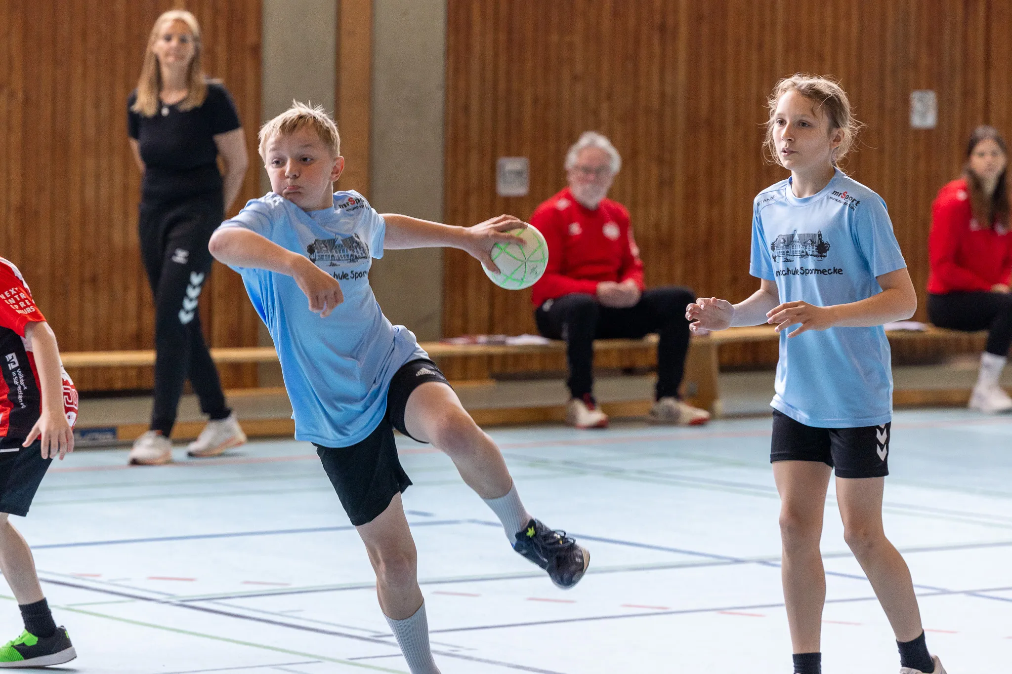 Zwei Kinder spielen Handball in einer Sporthalle. Der Junge wirft den Ball, während das Mädchen ihn abwehren will.