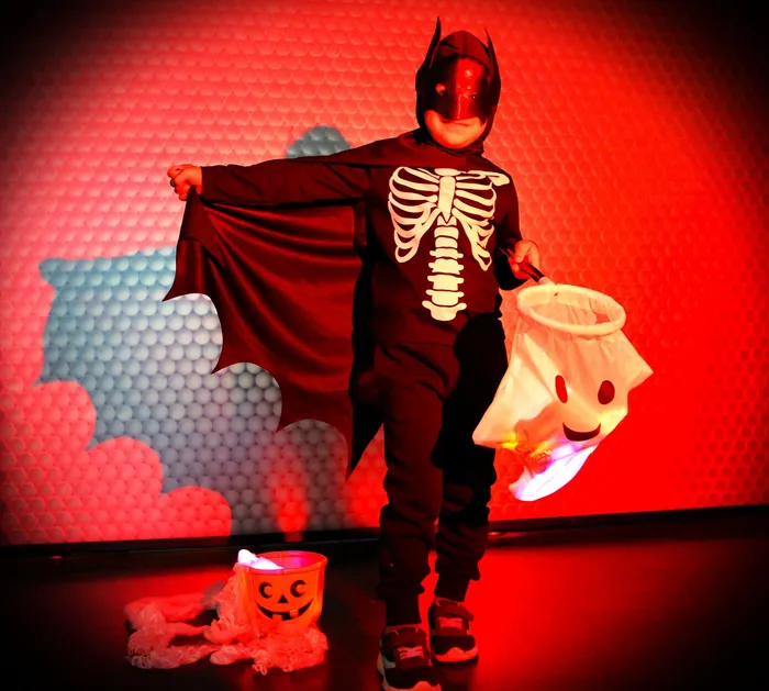 Ein Kind in einem Batman-Kostüm mit Skelett-Motiv steht mit einem leuchtenden Halloween-Eimer vor einem roten Hintergrund mit Ballons. Ein kleiner Kürbiskorb liegt vor dem Kind.