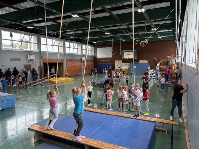 Kinder turnen in einer Turnhalle. Mehrere Kinder stehen Schlange, um an Seilen zu schwingen, während andere auf Matten und Geräten üben. Im Hintergrund stehen und sitzen weitere Kinder und Betreuer.