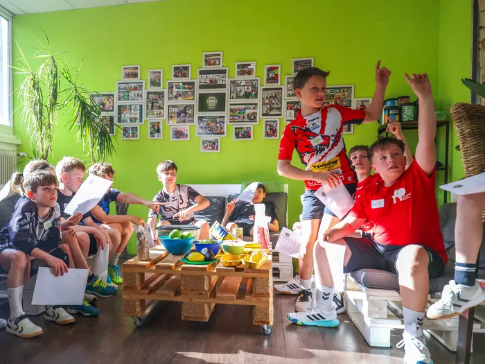 Eine Gruppe von Kindern sitzt und steht in einem Raum mit grüner Wand, einige halten Papier und Stifte. Zwei Jungen in roten Trikots stehen auf einem Sofa und heben die Arme. Auf der Wand hängen zahlreiche Fotos.