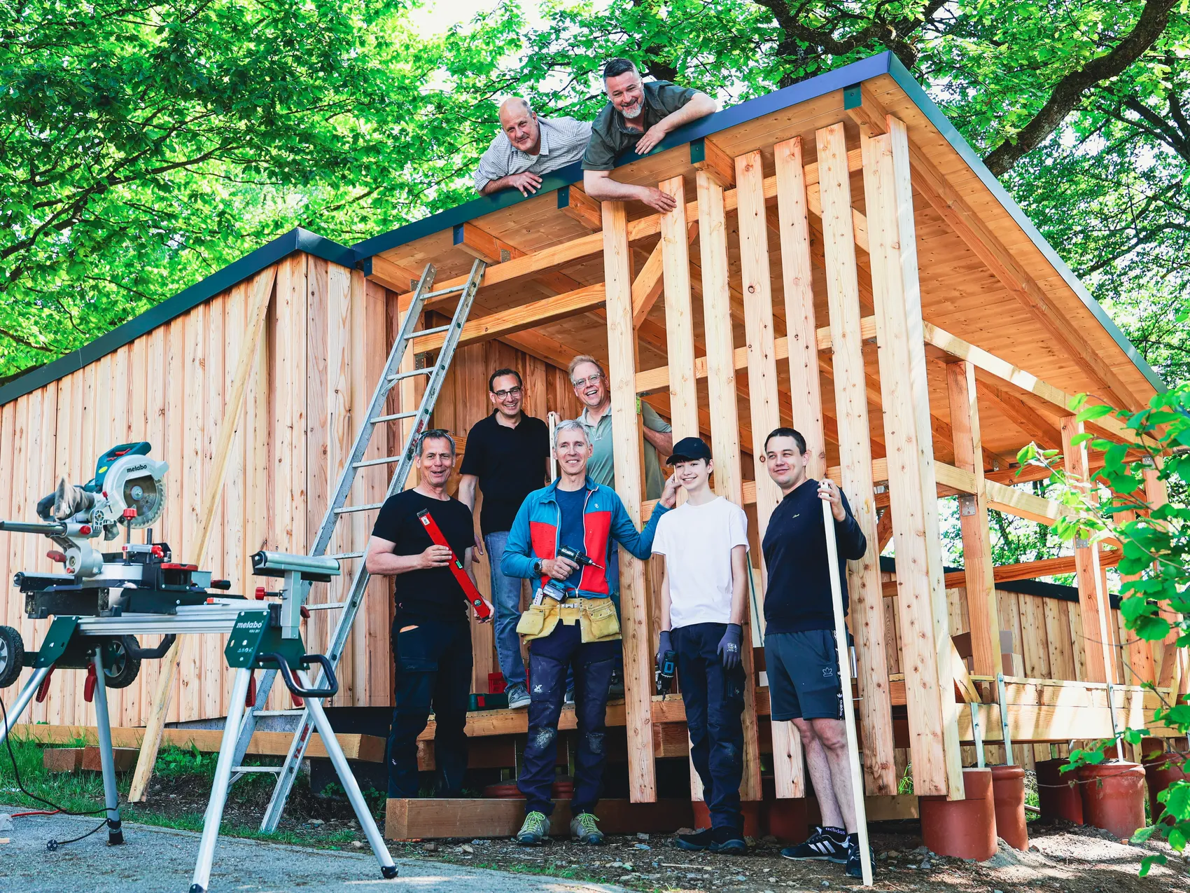 Eine Gruppe von sieben Personen steht vor und auf dem Dach eines Holzrahmens für eine Hütte, die im SOS-Kinderdorf gebaut wird. Einige halten Werkzeuge oder lehnen an Leitern.
