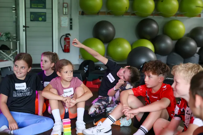 Eine Gruppe von Kindern sitzt im Kreis auf dem Boden und lauscht einem Vortragenden. Im Hintergrund sind Fitnessbälle und eine Tür zu sehen.