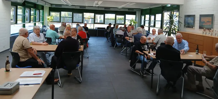 Mehrere Gruppen von Menschen sitzen an Tischen in einem hellen Raum mit großen Fensterfronten.