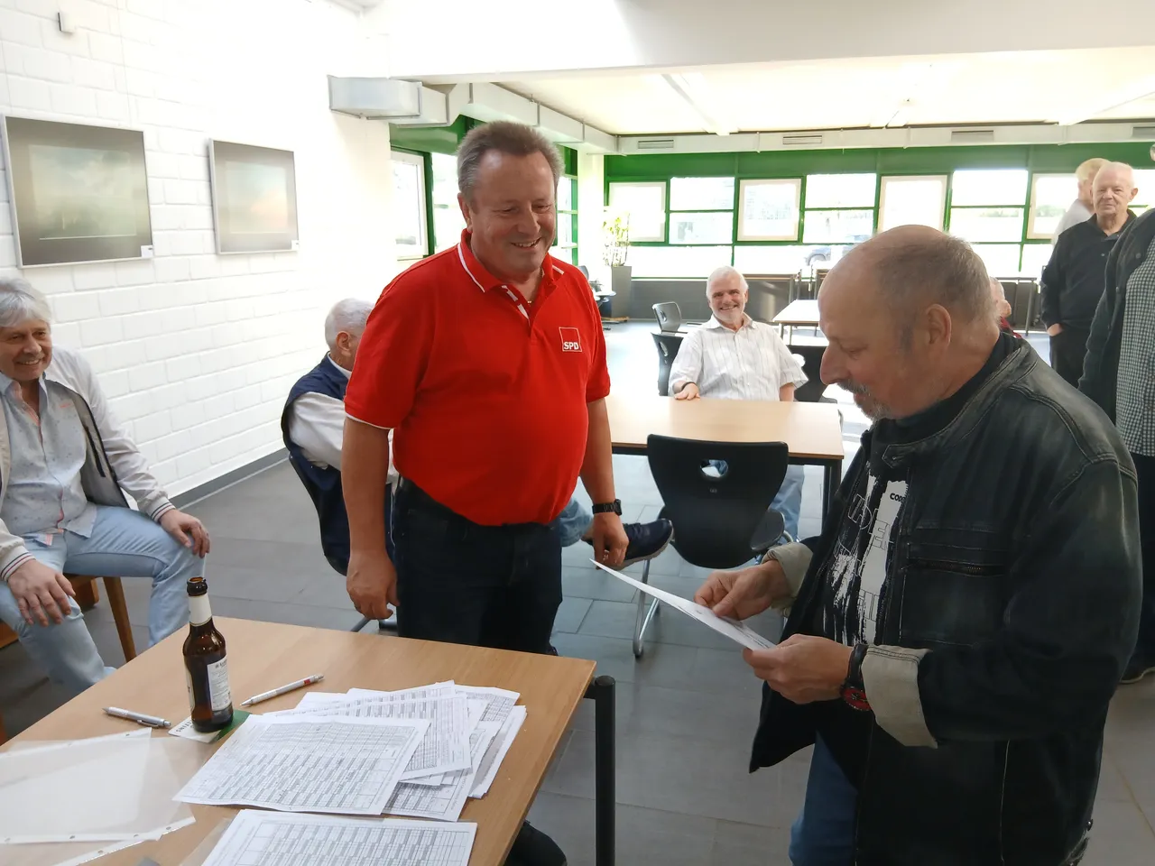Ein Mann im roten SPD-Poloshirt lächelt, während ein Mann in einer schwarzen Lederjacke ein Dokument betrachtet.