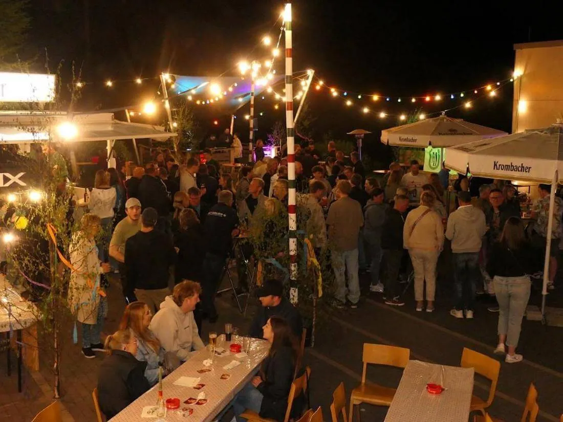 Menschen feiern im Freien, umgeben von Lichterketten und Schirmständern mit Werbeaufdruck. Im Hintergrund ein beleuchteter Stand und eine Menschenmenge.