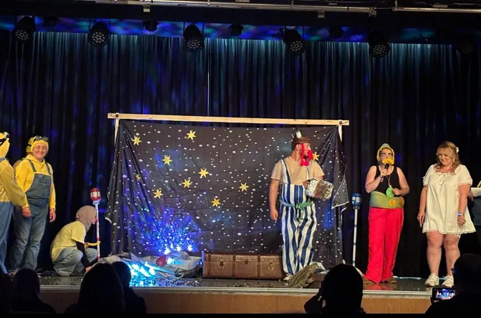 Bühnenbild mit Sternenhimmel und Requisiten. Darsteller in Kostümen, darunter ein Seemann mit Papagei, singen und spielen vor Publikum.