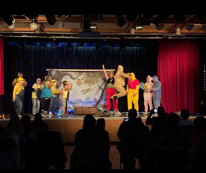 Kinder in Kostümen spielen auf einer Bühne das Theaterstück „Die besondere Reise durch die Wundertüte“. Im Hintergrund ein Bühnenbild mit einem Baum und Schriftzug „Hundred Acre Wood“. Das Publikum ist im Dunkeln zu erkennen.