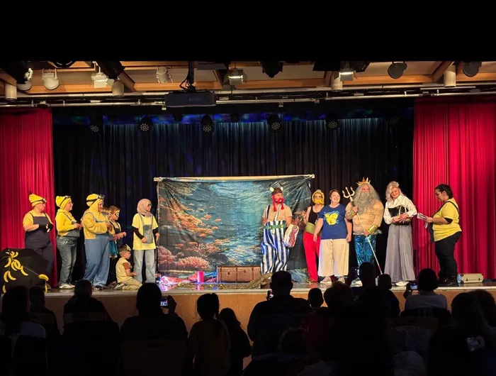 Kinder in Kostümen stehen auf einer Bühne vor einem Publikum und führen ein Theaterstück auf. Im Hintergrund ist ein Kulissenbild von Unterwasser zu sehen.