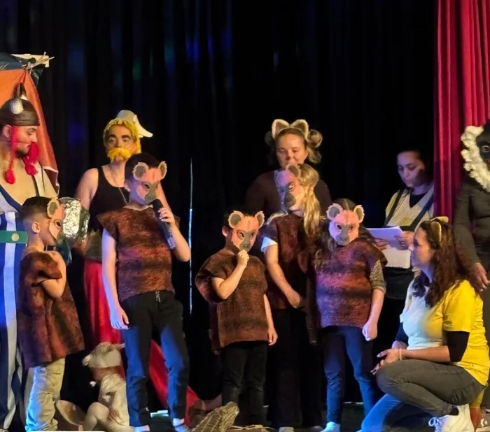 Eine Gruppe von Kindern und Erwachsenen in Tierkostümen – hauptsächlich Mäuse – steht auf einer Bühne und führt ein Theaterstück auf. Einige halten Mikrofone, andere Blätter mit Text. Im Hintergrund sind rote Vorhänge und weitere Personen zu sehen.