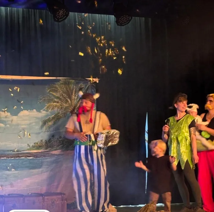 Ein Mann mit Piratenkostüm und -hut hält eine Schatzkiste vor sich, während Konfetti auf ihn herabfällt. Im Hintergrund stehen Kinder in Kostümen und ein Bühnenbild mit Palmen und Meer.