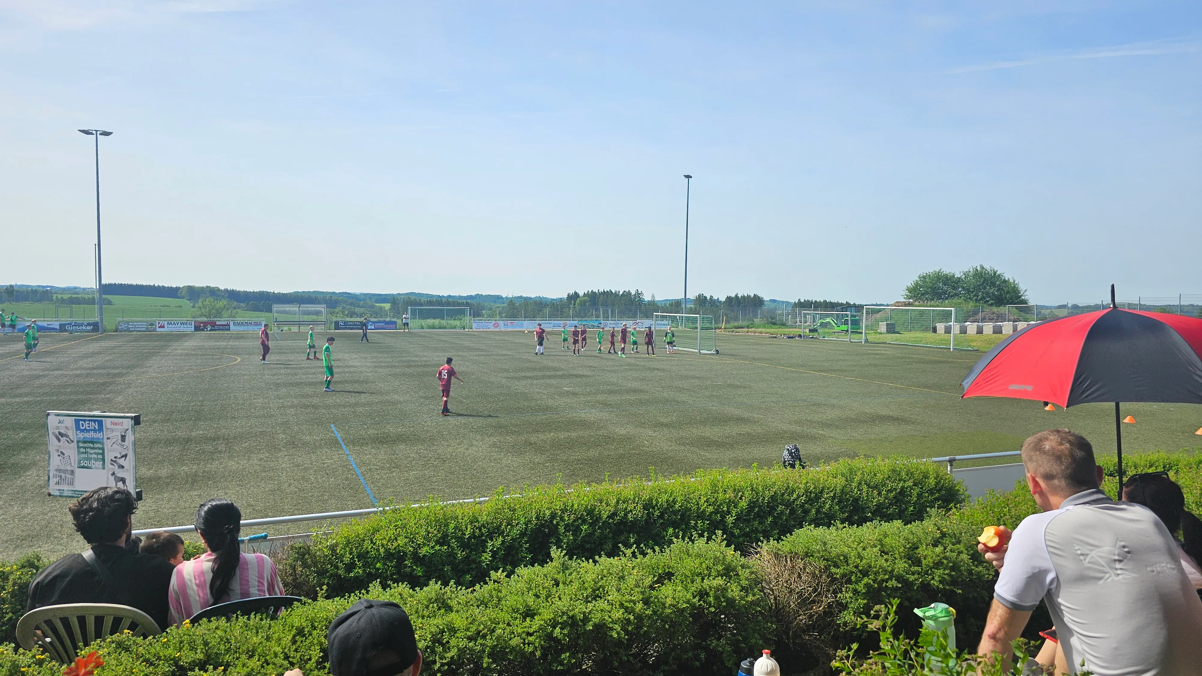 Fußballspiel auf einem Kunstrasenplatz mit Zuschauern am Spielfeldrand an einem sonnigen Tag.