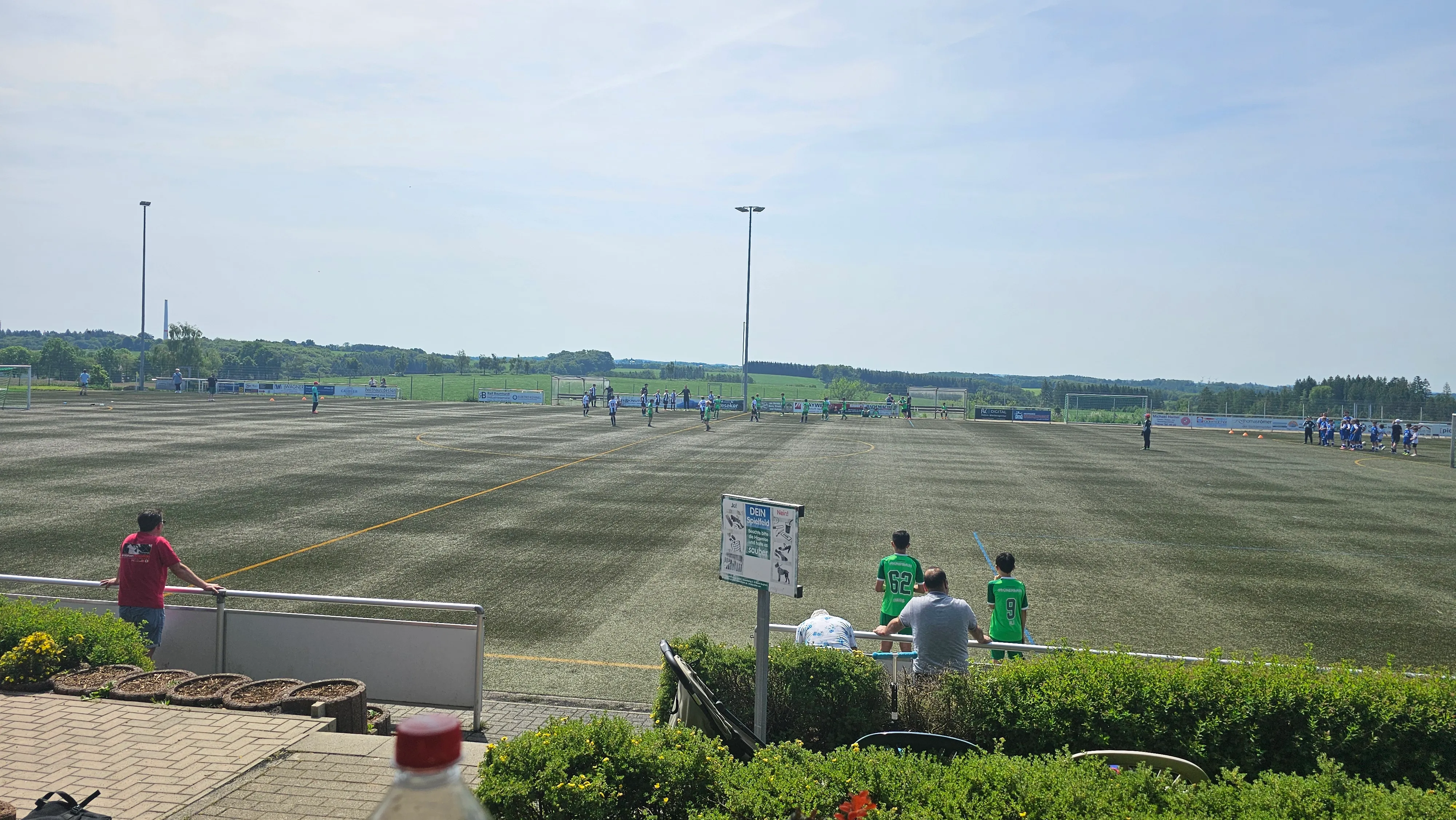 Fußballturnier auf einem Kunstrasenplatz mit mehreren Spielern und Zuschauern an einem sonnigen Tag.