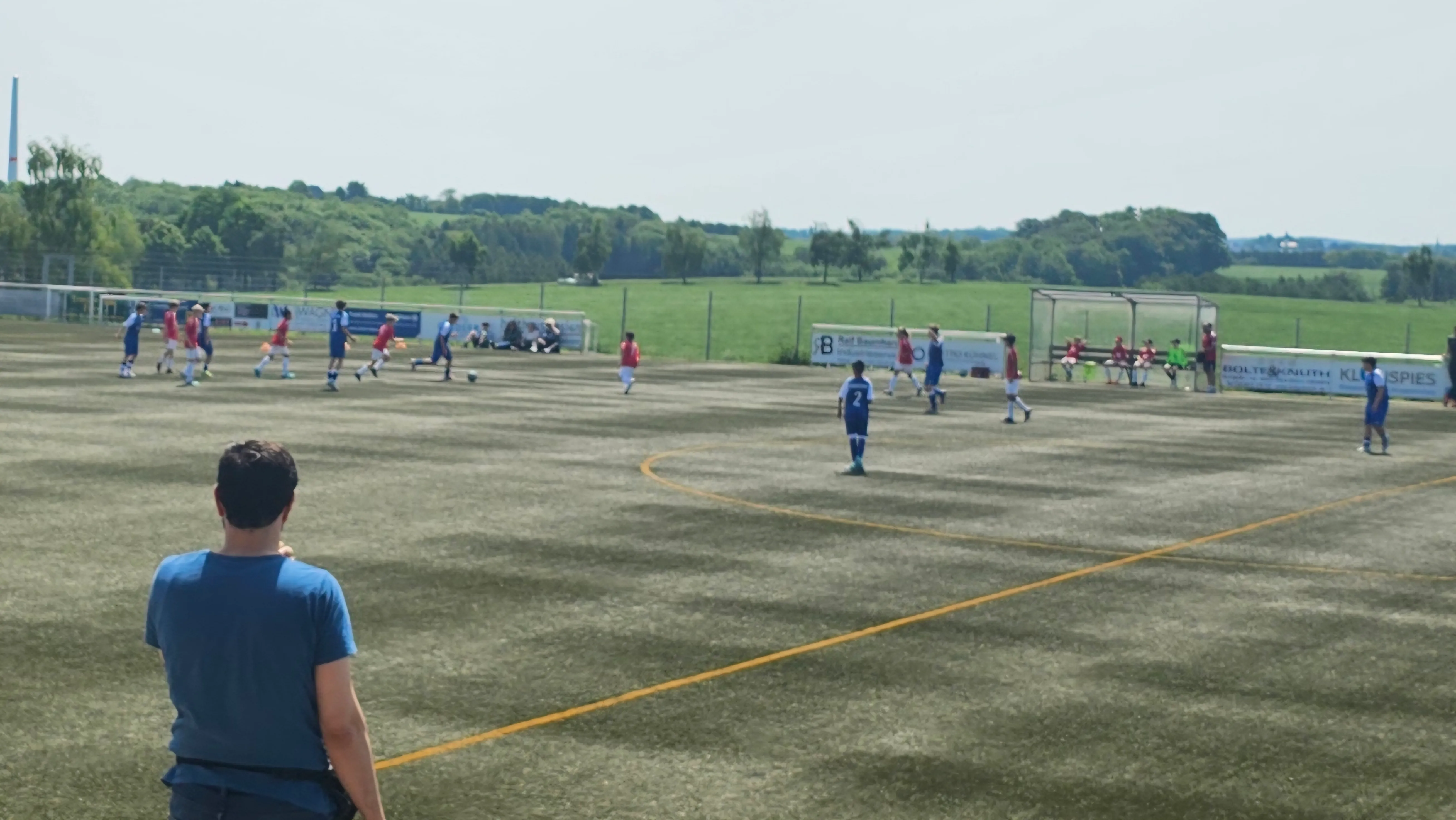 Fußballspiel auf einem Kunstrasenplatz mit zwei Mannschaften in blauen und roten Trikots. Im Hintergrund eine grüne Landschaft und Zuschauer am Spielfeldrand.
