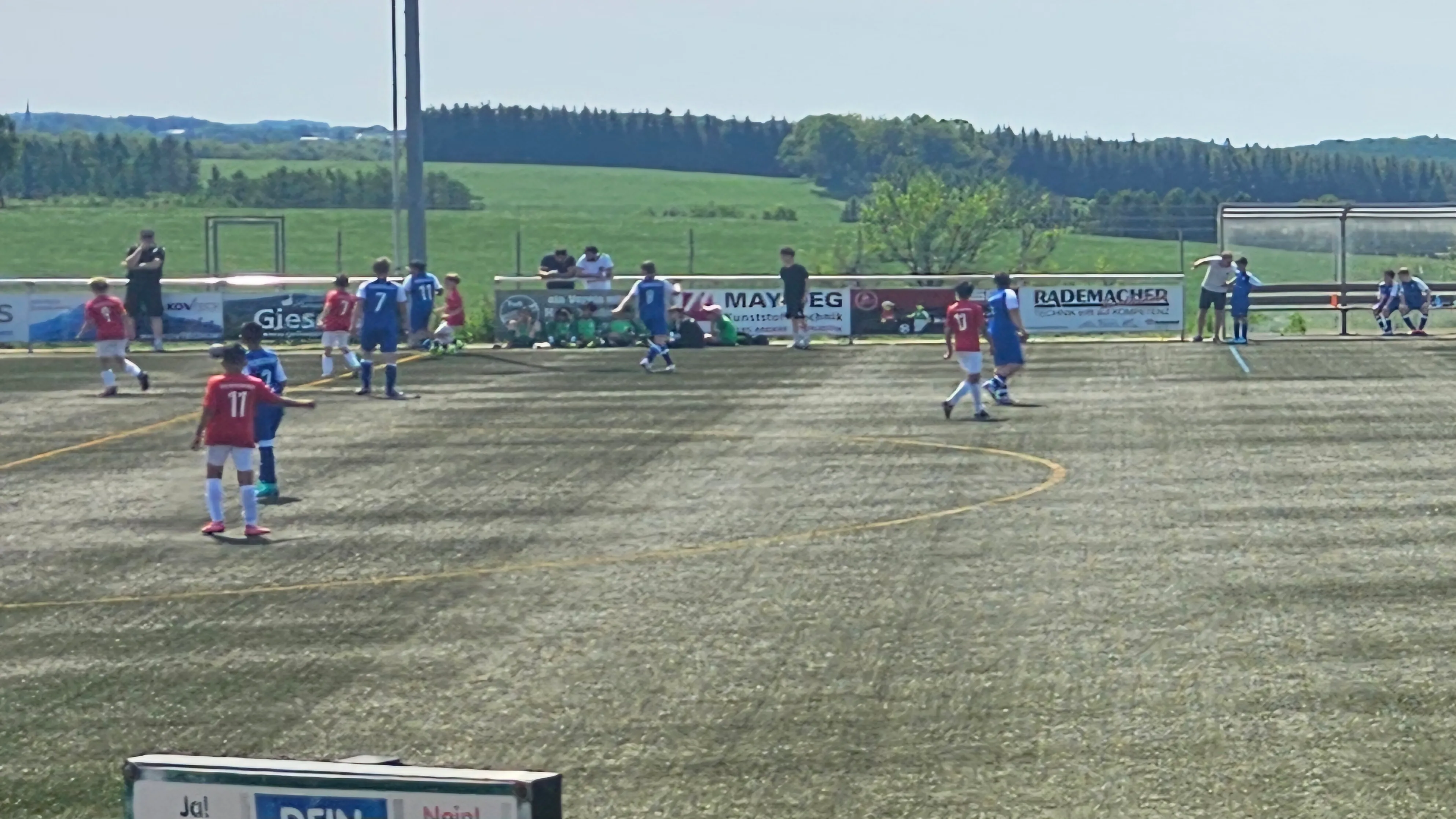 Fußballspieler in roten und blauen Trikots kämpfen um den Ball auf einem Kunstrasenplatz. Im Hintergrund eine grüne Hügellandschaft und Werbebanner.