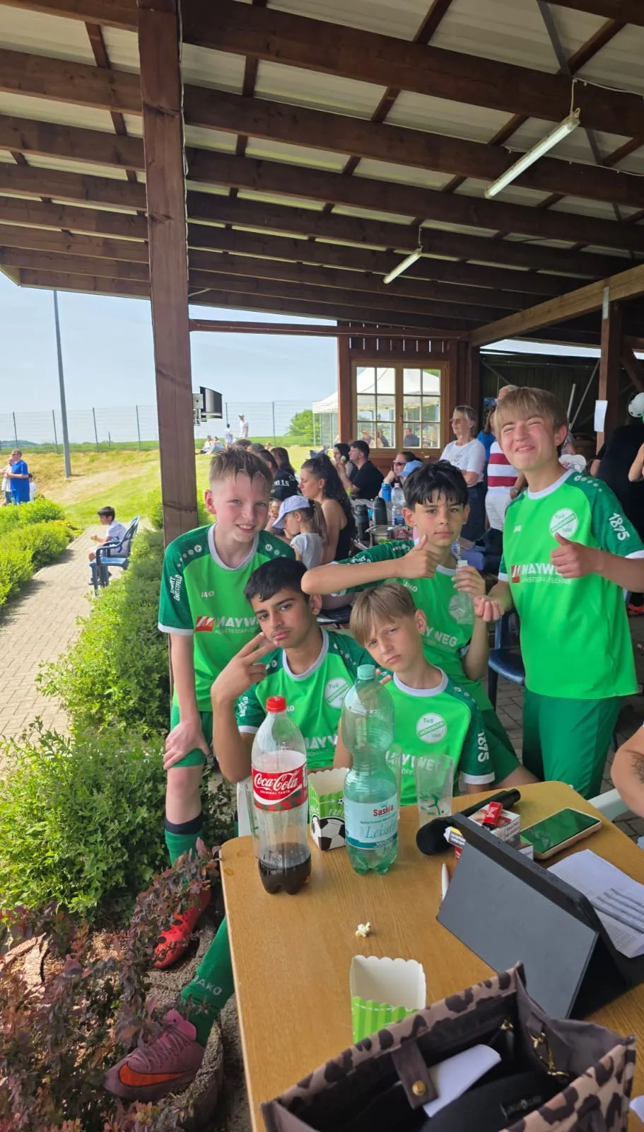 Fünf Fußballspieler im grünen Trikot des TuS Grünenbaum stehen an einem Tisch mit Getränken und Snacks unter einem Unterstand. Im Hintergrund sind weitere Personen und ein Fußballfeld zu sehen.