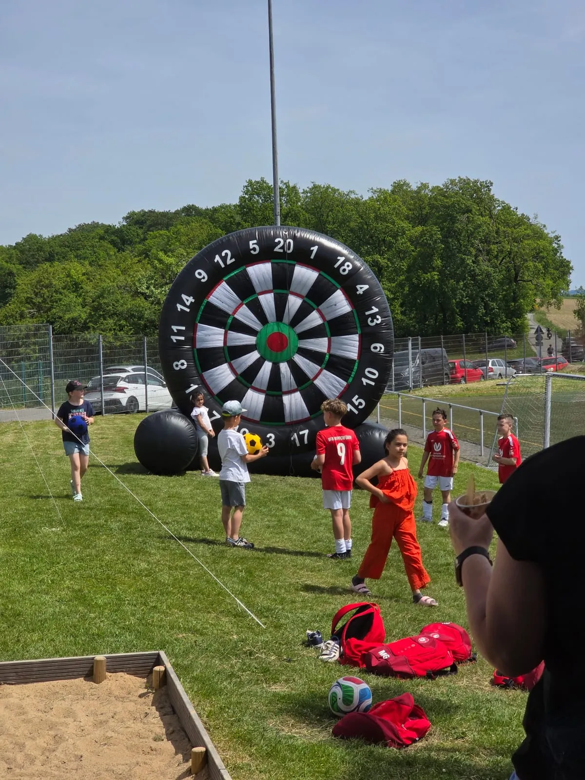 Kinder werfen Darts auf ein großes, aufblasbares Dartboard im Freien. Im Hintergrund eine grüne Landschaft und ein Fußballtor.