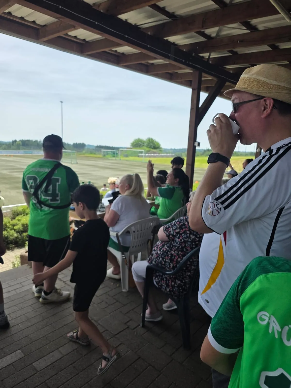 Zuschauer sitzen und stehen unter einem überdachten Bereich und beobachten ein Fußballspiel im Hintergrund. Einige tragen Sportkleidung, ein Mann trinkt aus einer Tasse.