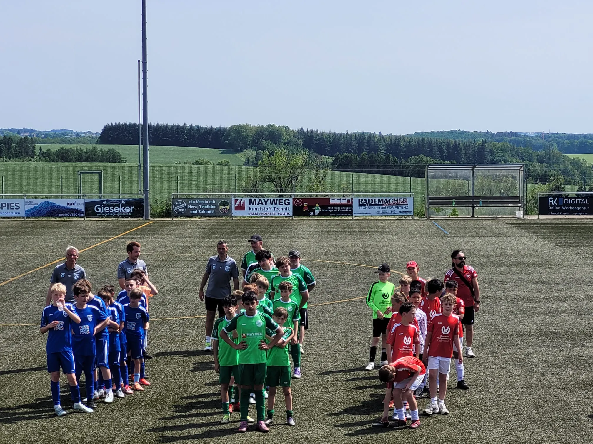 Zwei Jugendfußballmannschaften stehen sich auf einem Rasenplatz gegenüber, umgeben von Zuschauern und Sponsorenwerbung an einer Mauer im Hintergrund.
