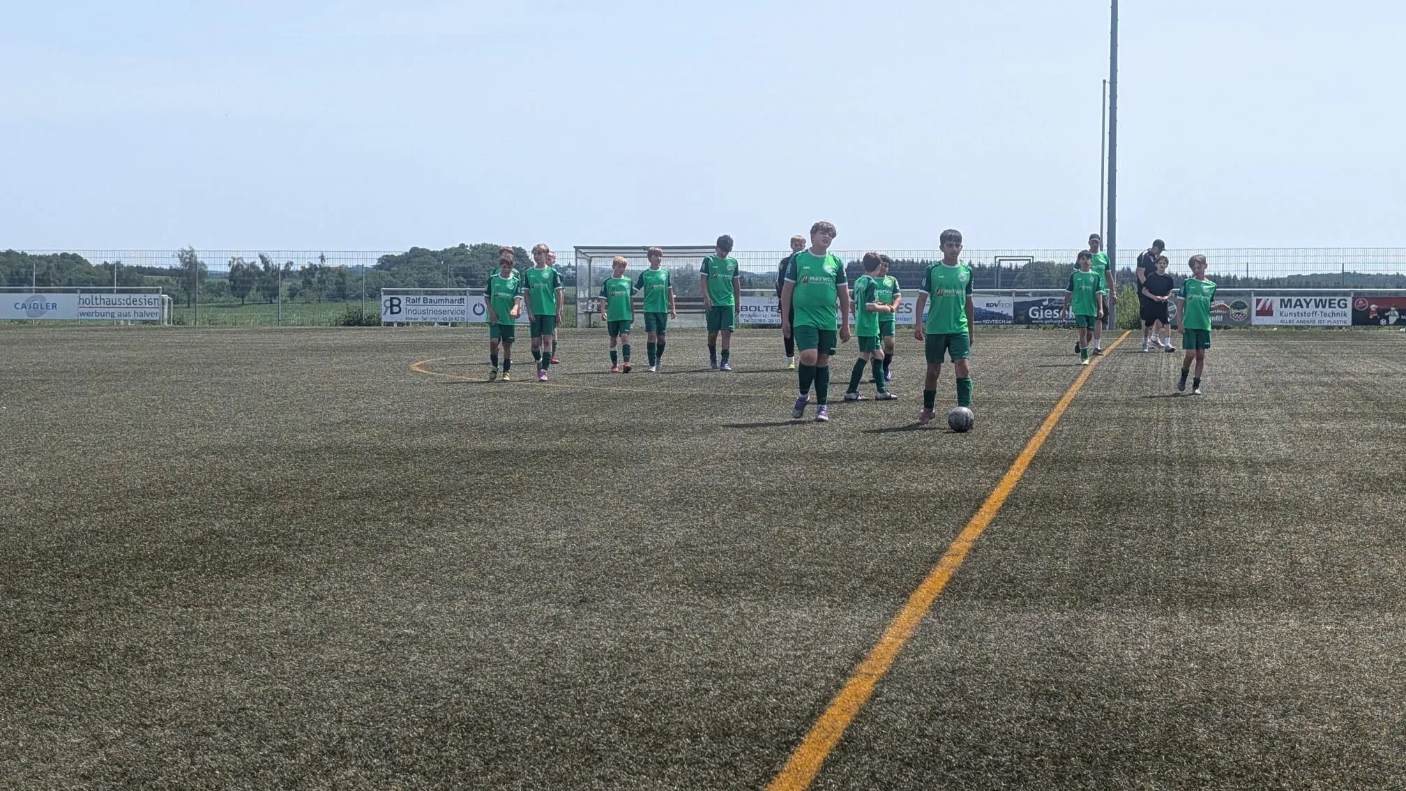 Fußballmannschaft in grünen Trikots steht auf dem Rasenfeld, bereit für ein Spiel. Im Hintergrund Werbebanner und Fußballtore.