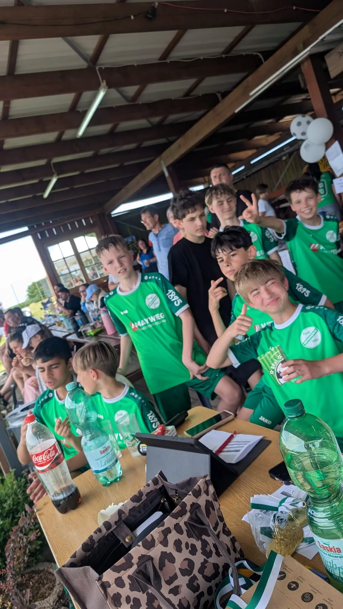 Eine Gruppe Jugendlicher im grünen Fußballtrikot mit der Aufschrift „1845“ sitzt und steht an einem Tisch unter einem überdachten Bereich. Auf dem Tisch stehen Getränke und Snacks.
