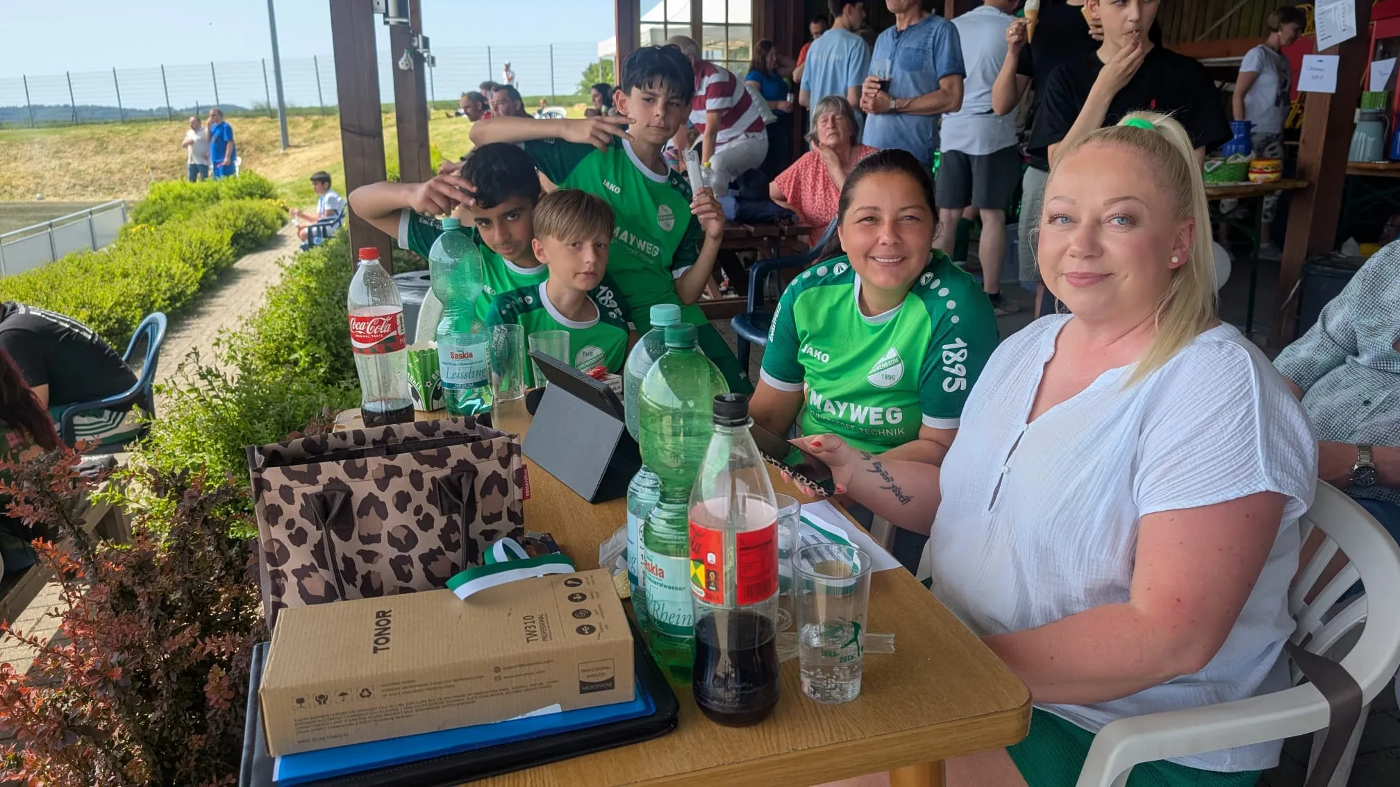 Gruppe von Menschen sitzt an einem Tisch im Freien, einige tragen Sportbekleidung mit dem Aufdruck „MAYWEG“. Im Hintergrund sind weitere Personen und ein Fußballfeld zu sehen.