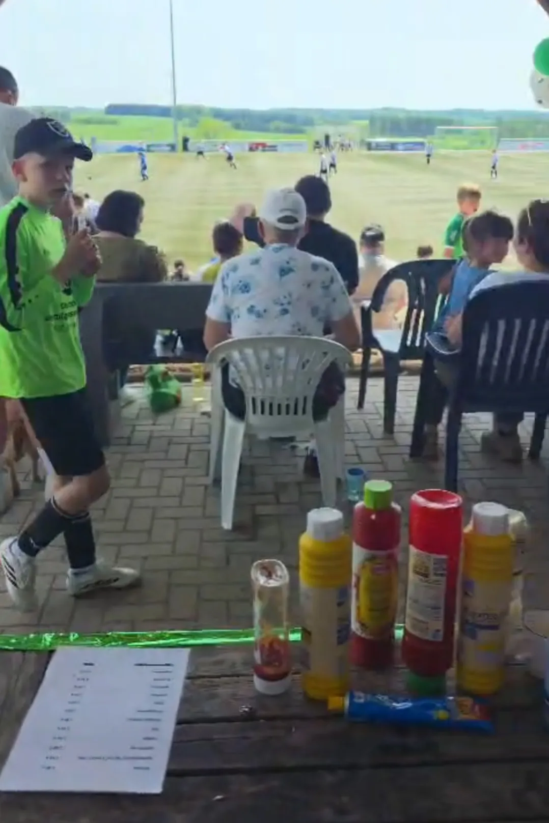 Zuschauer sitzen und stehen an einem sonnigen Tag an einem Fußballfeld. Im Vordergrund stehen Tische mit Essensutensilien und eine Ergebnisliste.