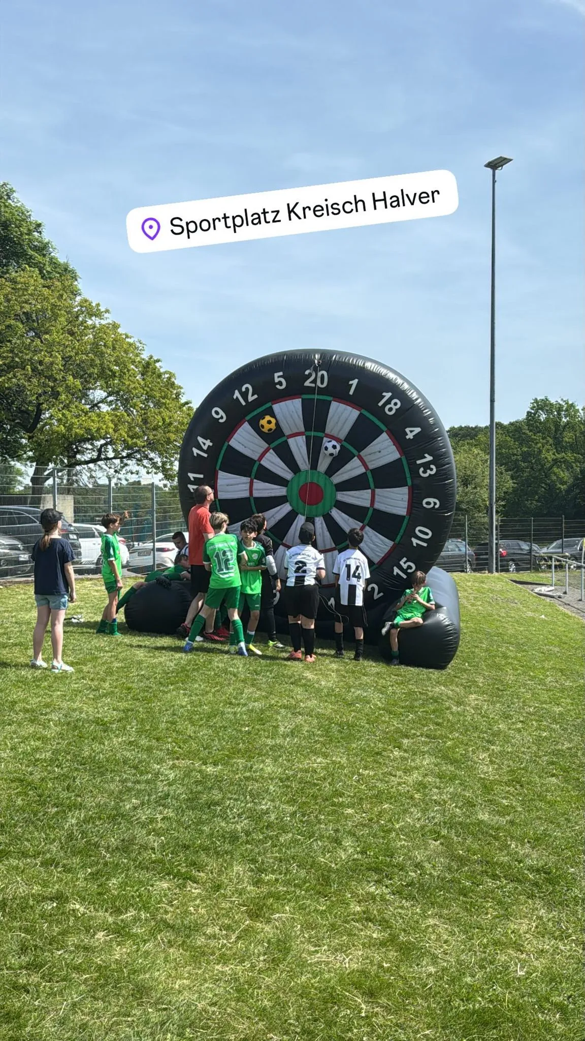 Auf einer Wiese steht ein aufblasbares Dartspiel mit den Zahlen 1 bis 20. Mehrere Personen in Sportkleidung stehen um das Dartspiel herum.