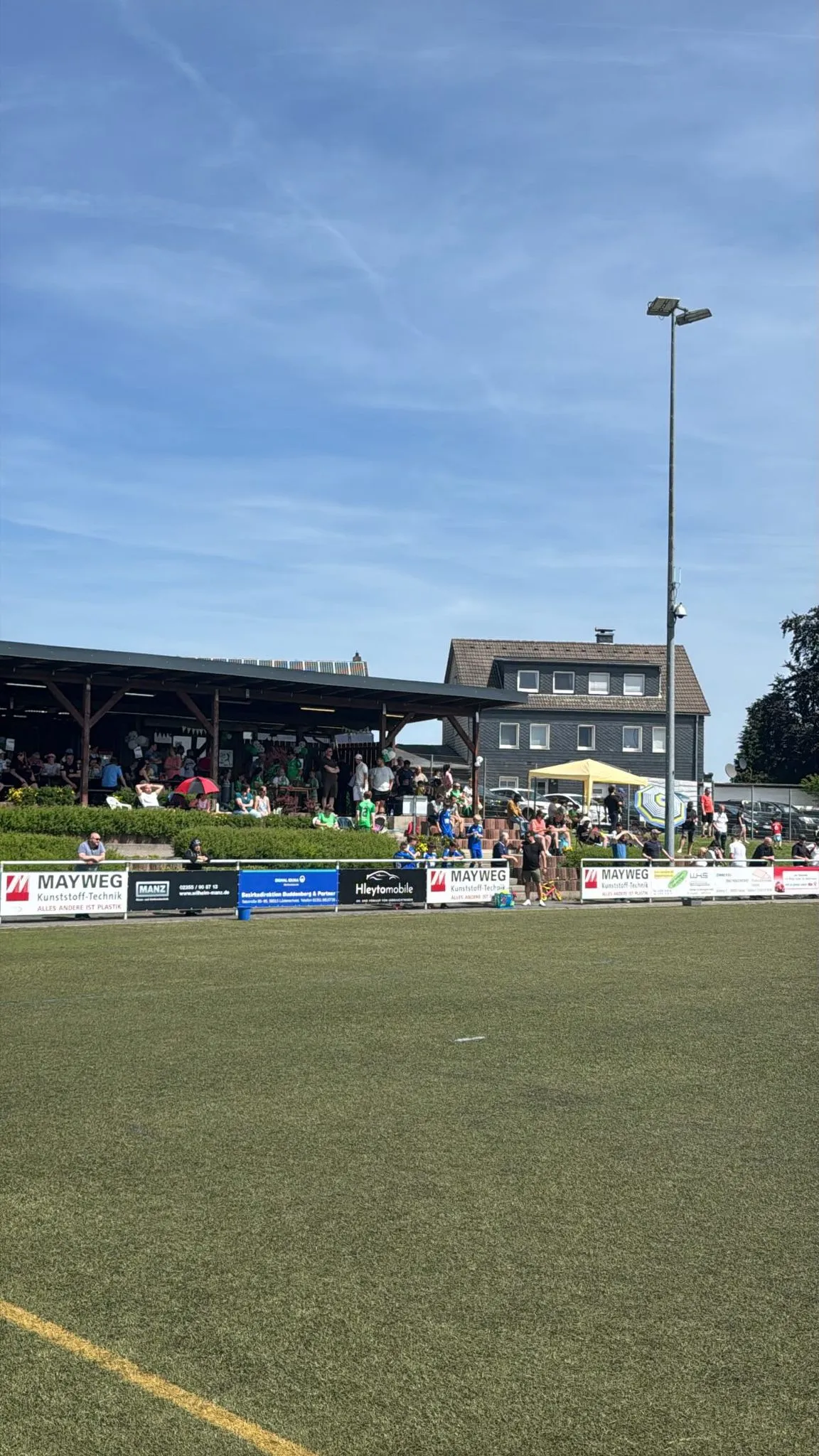 Fußballfeld mit Zuschauern unter einem überdachten Zuschauerstand und Werbebanden am Spielfeldrand an einem sonnigen Tag.