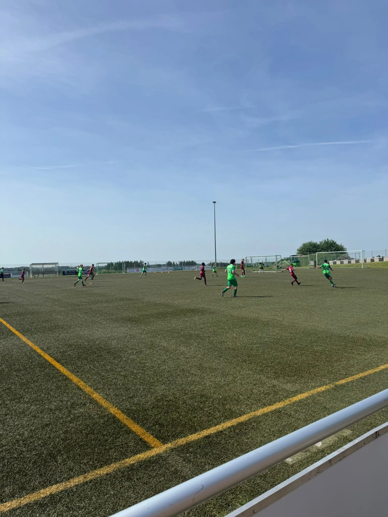 Fußballspieler in grünen Trikots auf einem Rasenplatz, im Hintergrund ein Mast und Bäume unter blauem Himmel.
