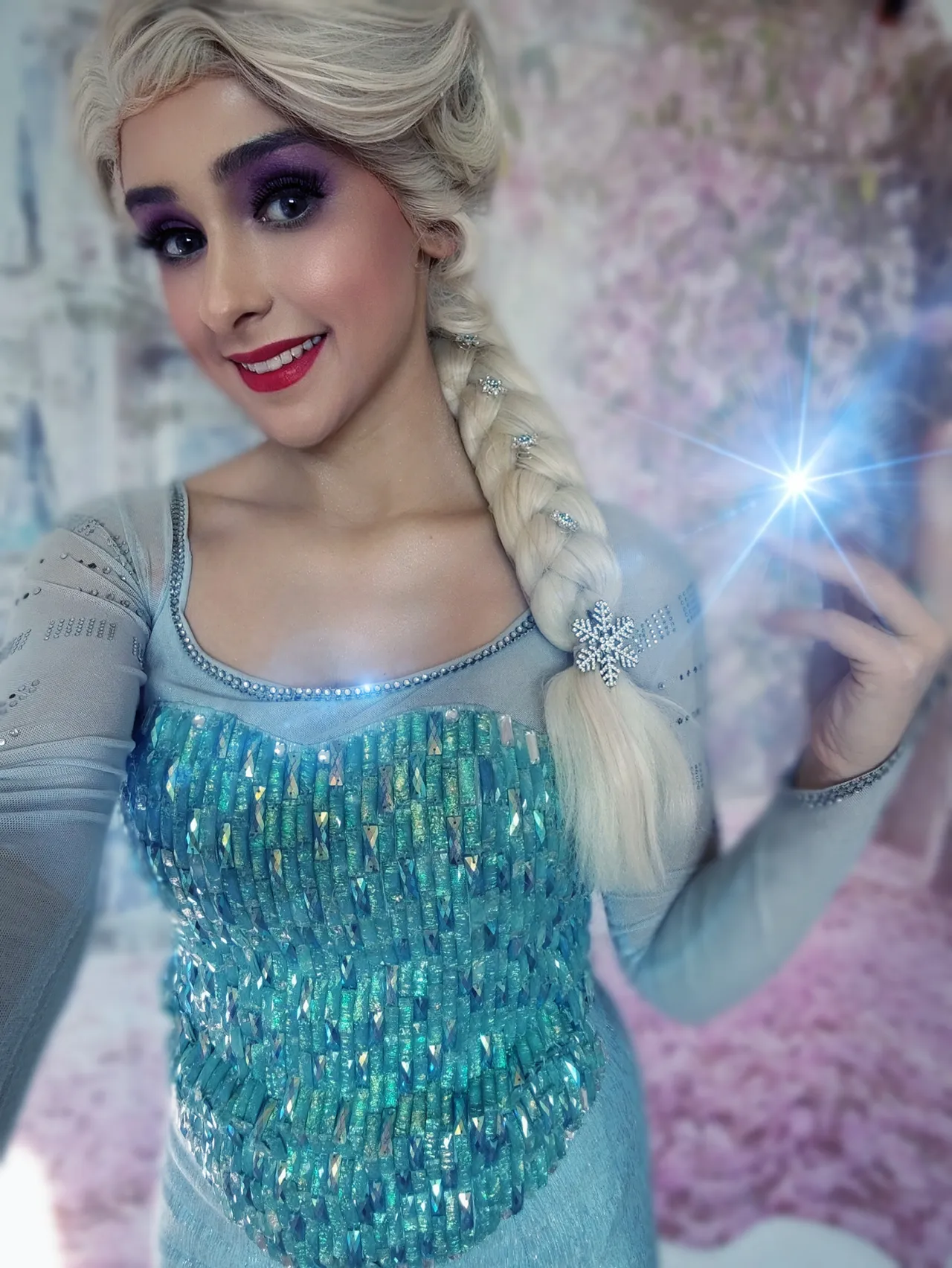 Eine Person mit blondem, geflochtenem Haar und in einem eisblauen Kleid mit Pailletten posiert lächelnd. Sie hält eine Hand mit einem leuchtenden, blauen Lichteffekt.