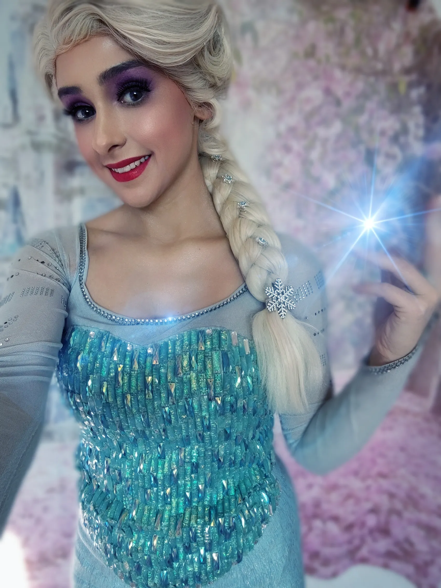 Eine Person mit blondem, geflochtenem Haar und in einem eisblauen Kleid mit Pailletten posiert lächelnd. Sie hält eine Hand mit einem leuchtenden, blauen Lichteffekt.
