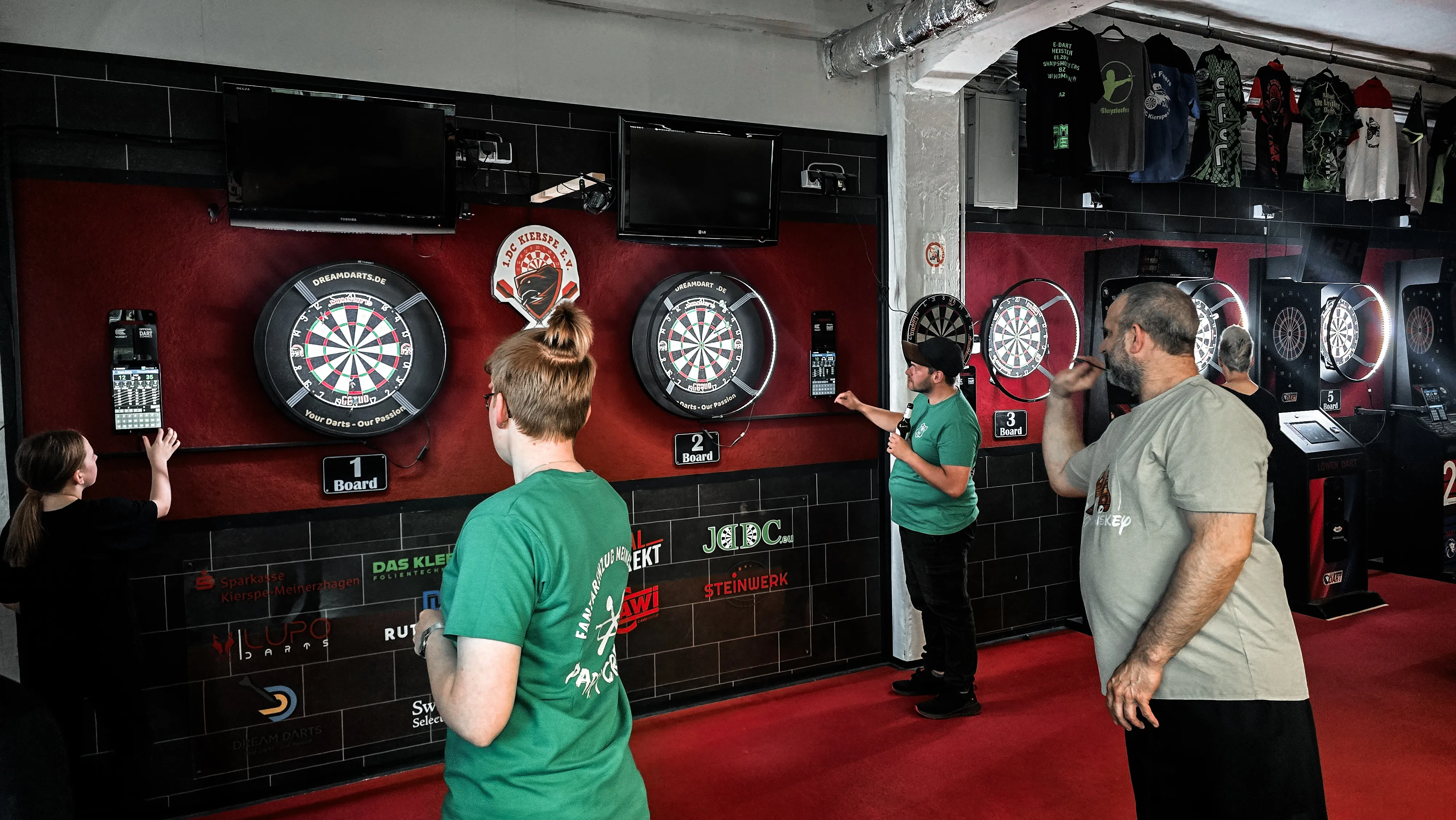 Drei Dartscheiben an einer dunklen Wand, vor der ein Mädchen, ein Mann und eine Frau stehen und sich dem Spiel zuwenden.