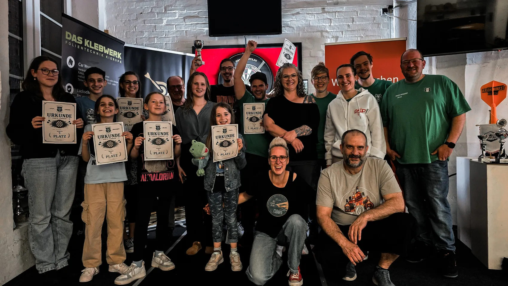 Gruppe von Menschen hält Urkunden und Pokale in einem Raum mit Bannerwand, darunter Mitglieder des Fanfarenzugs Meinerzhagen und des Dartclubs Kierspe.
